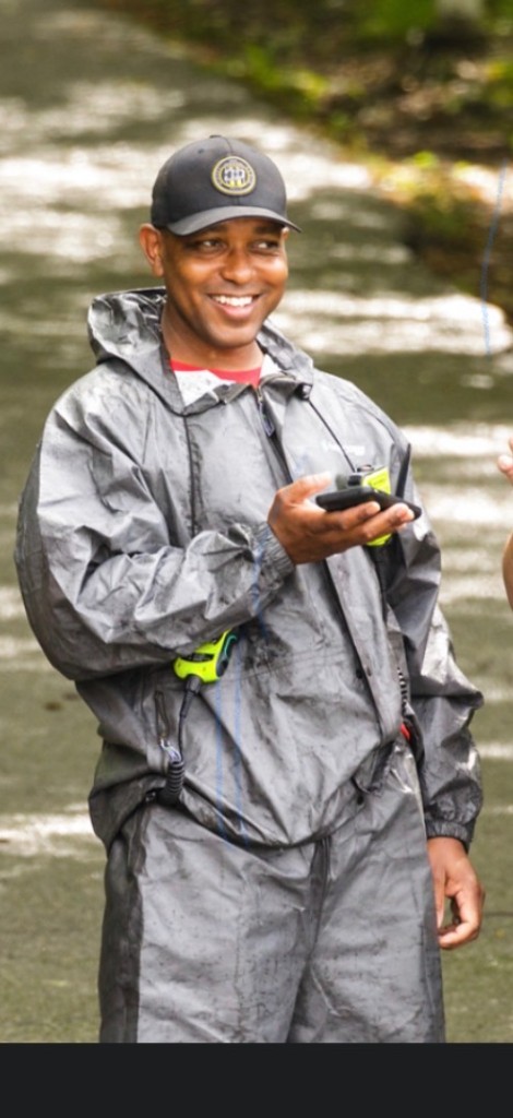 Reginald Wadley outdoors in grey protective rain gear, smiling and holding a walkie-talkie, with a second radio clipped at his waist
